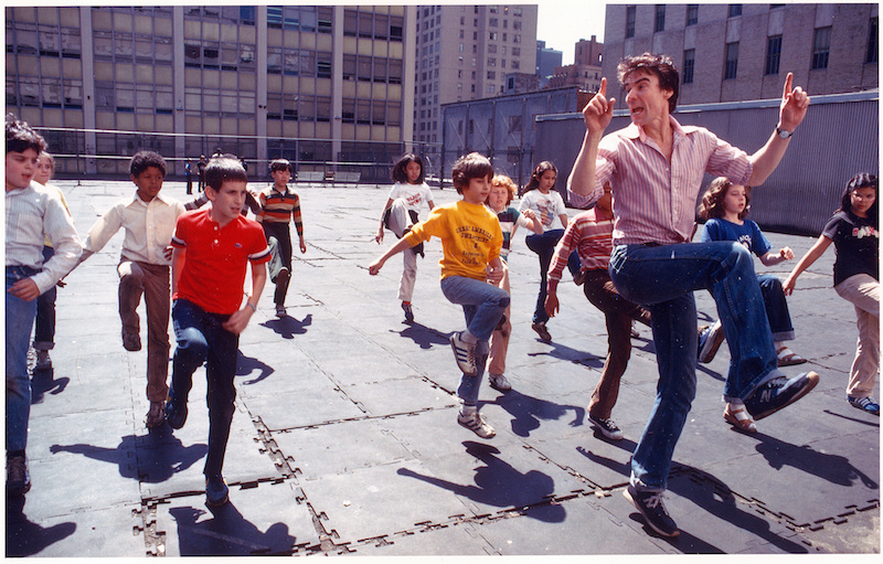 Jacques leads a group of kids on the roof of their school. Their knees are hiked into the air.